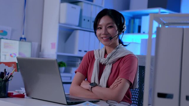 Smiling happy Asian woman customer service agent working late night overtime wearing headset laptop modern office call support