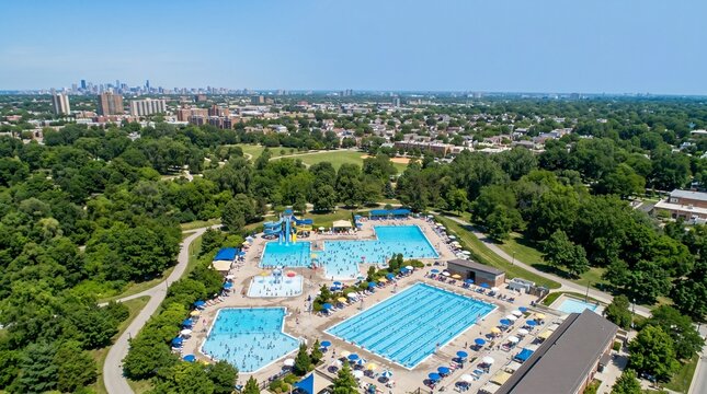 An aerial view of a sprawling public swimming pool complex on a sunny day. The complex features multiple pools, including a large lap pool and a recreation pool with slides.