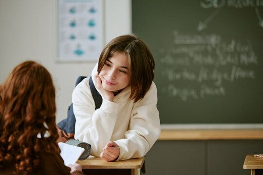Teenage Caucasian schoolgirl leaning on desk, listening to classmate in classroom, showing peer communication and school engagement