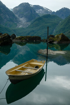 A small rowboat floating on the crystal-clear waters of Bondhusvatnet beneath the dramatic mountains and glaciers of western Norway. The image captures the calm atmosphere, mirror reflections, and unt