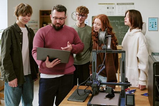 Medium shot of middle aged Caucasian male teacher guiding teenagers using laptop beside 3D printer in classroom