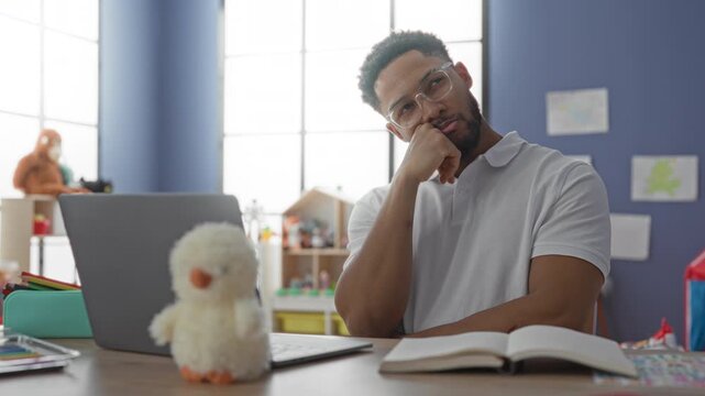 Man typing on laptop with hand to chin beside stuffed toy and open book in classroom building; concentration.