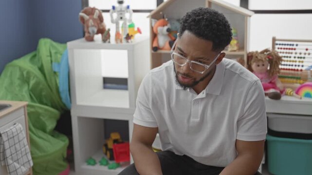 Man gesturing hand toward an abacus and toys while seated in a playroom inside a building wearing glasses and a white polo; guidance learning thoughtful.