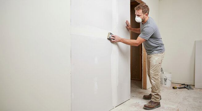 Male contractor sanding drywall seam in unfinished room. Construction worker wearing dust mask smoothing plaster wall during interior home renovation