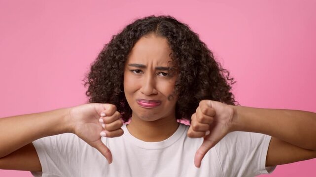 A young woman with curly hair expresses disappointment by giving a thumbs down gesture. She has a neutral shirt on and appears to be in a studio setting with pink background.