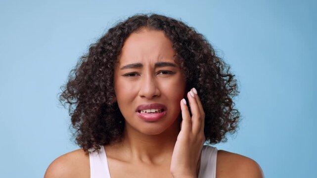 A woman with curly hair is seen with a pained expression on her face. She is holding her jaw with her hand, indicating she is feeling discomfort. The background is light blue.