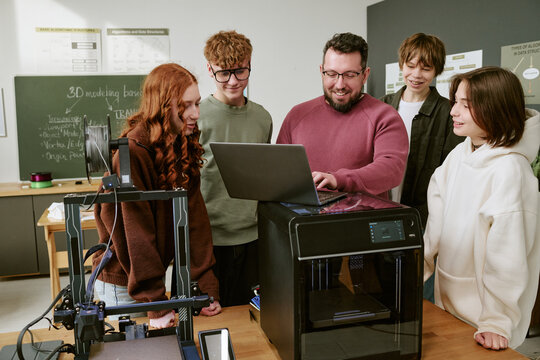 Teenagers with male teacher reviewing laptop beside 3D printers in classroom, learning digital fabrication and STEM skills