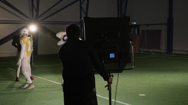 Medium full shot of Caucasian male television production gaffer setting up lights at sports arena to film show, while American football players are adjusting protective breastplates before game