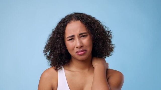 A young African American woman stands against a solid blue background. She looks uncomfortable while holding her neck, expressing discomfort. Her curly hair frames her face as she reacts.