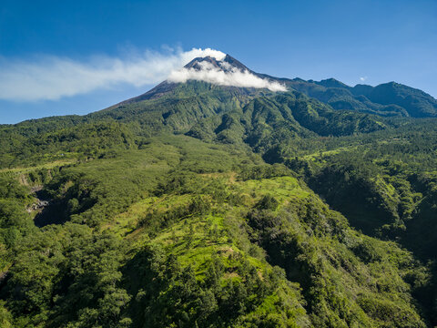 Aerial view of Mount Merapi volcano with smoke rising from the crater and lush green slopes under a clear blue sky in Kecamatan Cangkringan, Daerah Istimewa Yogyakarta, Indonesia.