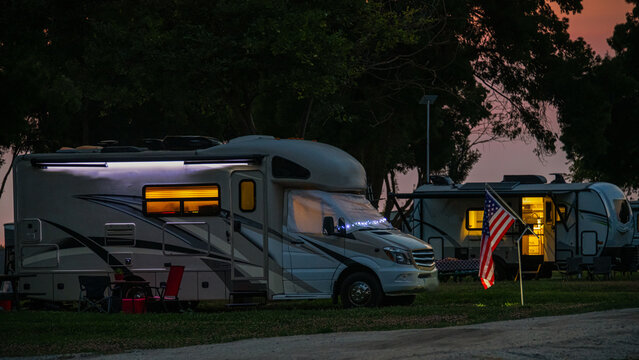 Two RVs are parked at a campsite during sunset, with lights glowing warmly inside. An American flag waves in the gentle evening breeze.