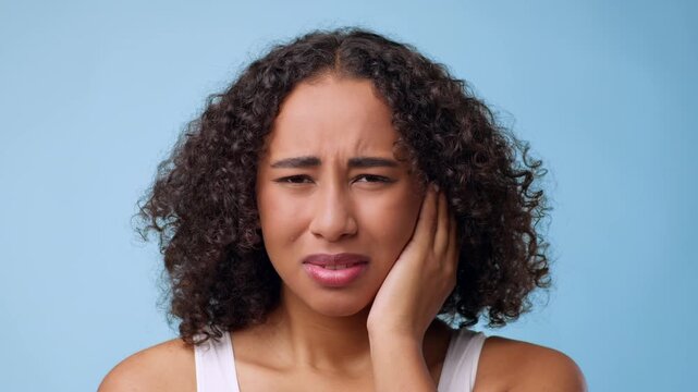 A woman with curly hair is showing an expression of discomfort. She is standing against a blue background while touching her face. The scene captures a moment of concern or pain.