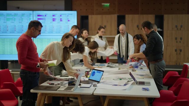 A group of colleagues actively collaborates around a table in a modern office space. Businesspeople engage in a focused discussion, analyzing documents and digital data on laptops.