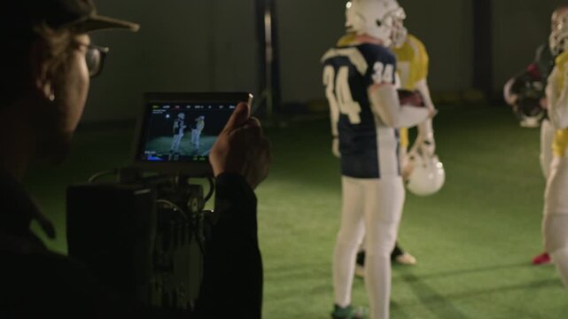Over-shoulder shot of Caucasian male cinematographer walking up to professional video camera on tripod and setting up frame to film gridiron football players train on pitch for documentary