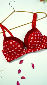 Close up of a red women's bra with white flower patterns hanging on a golden hanger with scattered pink petals lingerie underwear fashion woman clothing lingerie underwear fashion woman clothing