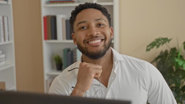 Man holding eyeglasses by chin, smiling at computer desk with bookshelf and houseplant in building; confidence warmth.