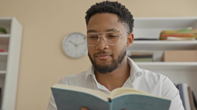 Man reading a paperback book with glasses, holding open pages with shelves and wall clock visible in a building study space; learning concentration calm.