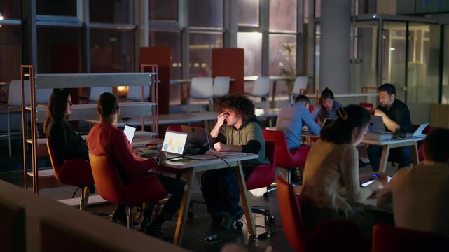 A diverse group of professionals are diligently working on their laptops in a modern office environment late at night. The team collaborates and focuses on their projects during extended hours.