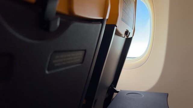 Airplane cabin view with seat tray, window, and soft daylight during flight. Quiet travel moment from inside the aircraft before or after takeoff. Horizontal video on a plane.