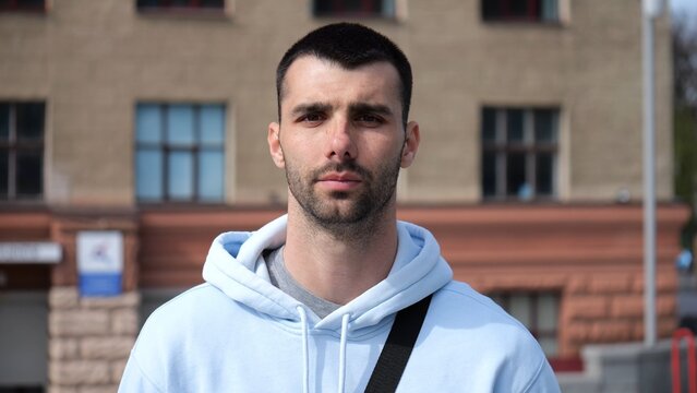 Portrait of young confident bearded man in casual clothing with cityscape background. Close up of handsome guy looks into camera standing at city environment at sunny day