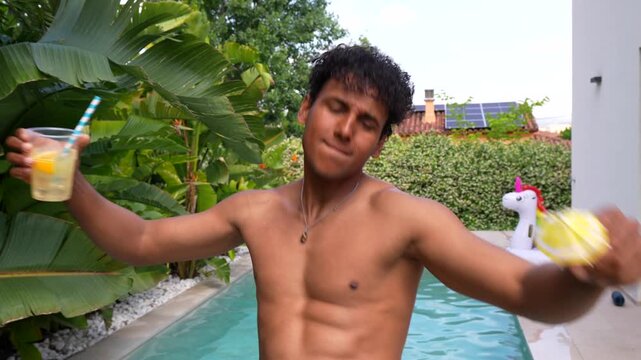 Charismatic dark skinned man with an athletic body filming himself while dancing and drinking a cocktail in a swimming pool, celebrating the beginning of summer holidays at a resort