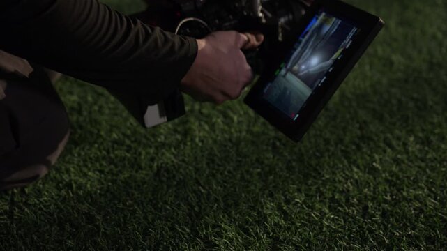 Medium close-up of hands of anonymous cinematographer filming gridiron football players performing ball kickoff at match at ground level, crouching above green turf, scene visible on camera viewfinder