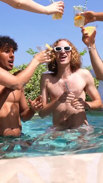 Happy diverse group of young friends dancing and toasting with cocktails in a swimming pool on a sunny day, enjoying their vacation at a villa with an inflatable unicorn in the background