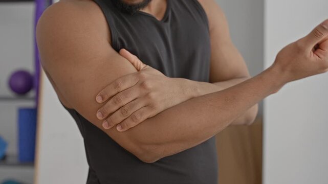 Man massaging bicep and holding forearm after workout in a fitness studio with foam roller and exercise ball visible; recovery relief focus.