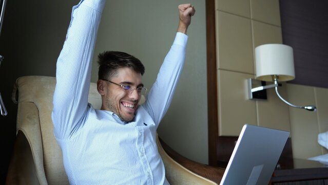 Young businessman working on laptop in modern hotel room and reacting to good news with excitement and happiness. Happy man celebrating success while using computer show achievement and motivation