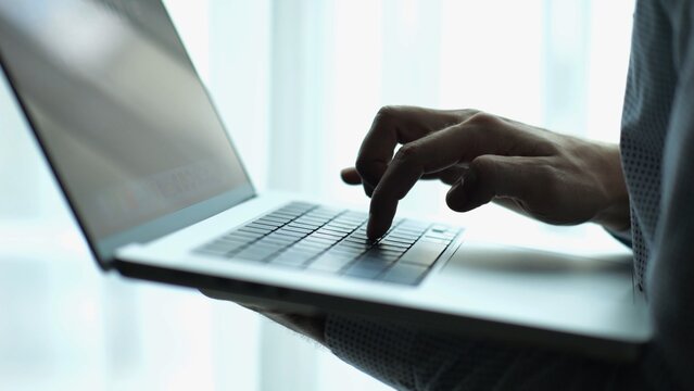 Male hand of young businessman typing text on laptop keyboard near window in modern office. Professional manager working on computer with natural light, showing productivity and communication