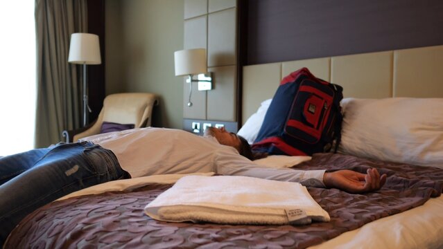 Exhausted man lying on bed in modern hotel room after long business trip. Tired young businessman relaxing in comfortable accommodation interior after work, travel and stressful daily routine
