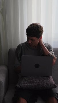 Neck pain. Man on couch with laptop rubbing neck in discomfort, slouched posture and tilted head, pillow on lap, soft curtain backlight, remote worker or freelancer, vertical