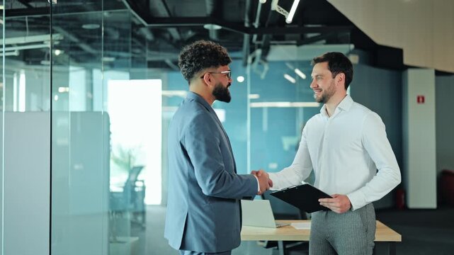 Medium shot of Middle Eastern businessman and Caucasian colleague shaking hands in office. Diverse partners greeting each other before meeting or celebrating successful partnership agreement.