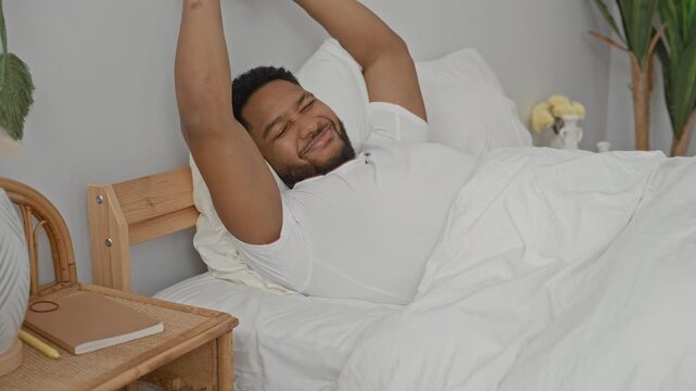 Man stretches arms overhead in bed against a wooden headboard in a building, smiling with eyes closed and relaxed posture; calm contentment.