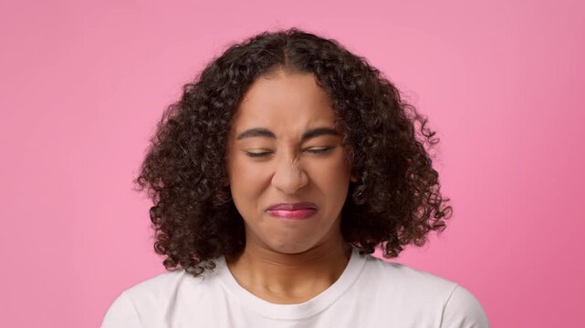 A young African American woman with curly hair squints her eyes and makes a playful face. She stands against a bright pink backdrop, expressing joy and humor.