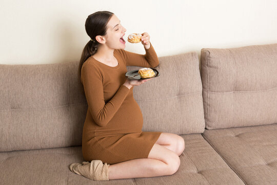 Young pregnant woman enjoys eating tasty cake resting on the sofa at home. Unhealthy diet during pregnancy concept