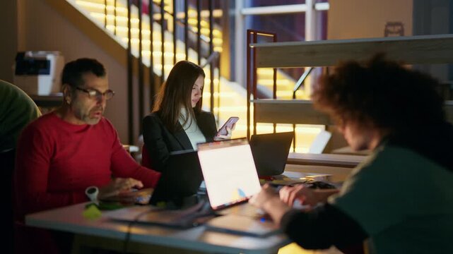 A dedicated business team works diligently on laptops in a modern office environment late at night. Colleagues are focused on their tasks, illustrating commitment and productivity after hours.