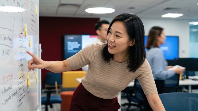 Young woman pointing at whiteboard during collaborative office brainstorming session, smiling and engaged in planning with colleagues