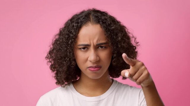 A young woman with curly hair stands against a pink wall. She shows an intense expression, pointing her finger as if making a serious point. Her look suggests urgency or concern.