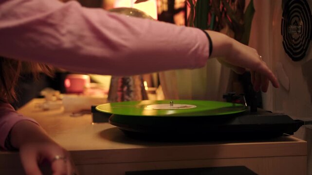Closeup of oldfashioned record player with warm ambient lighting. Green vinyl record spins in cozy retro decor ambiance. Closeup of female hand puts the needle of turntable in and turns on the music