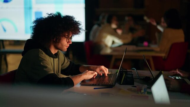 A focused young man with curly hair and glasses works diligently on his laptop in a dimly lit office. He is actively typing while colleagues work in the background, with data projected on a screen.