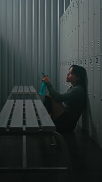 Young Sporty Woman Sitting on Floor, Leaning Against Lockers in Dressing Room. Girl Tired and Stares Blankly, Reflecting on Training. Burnout, Mental Challenge of Professional Sports. Vertical Shot.