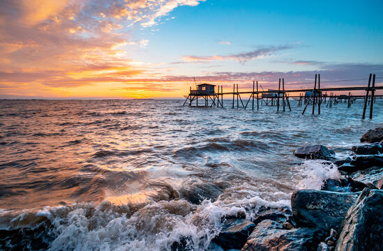 Cabane de p&ecirc;cheurs sur pilotis vue au coucher du soleil. Vagues et mousse sur les roches sombres au premier plan, pr&egrave;s d'Yves en Charente-Maritime, France