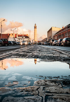 Calais, le phare du port de vu depuis le sol pav&eacute; de la rue se refl&eacute;tant dans une flaque d'eau, tandis que des voitures circulent dans la rue. Moment captur&eacute; dans le nord de la France.