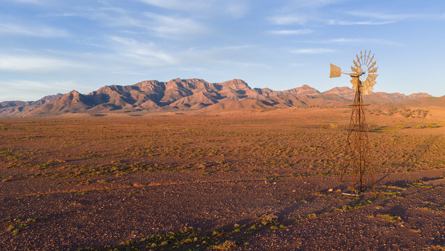 Aerial view of a traditional windmill standing in the arid outback landscape with rugged mountain peaks under a clear sky Flinders Ranges, South Australia, Australia.
