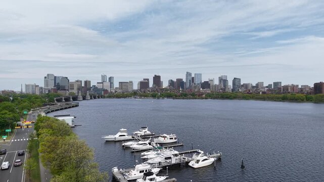 Aerial drone view looking down the Charles River in Boston, featuring luxury boats docked at a marina with the historic Longfellow Bridge, Storrow Drive traffic, and city skyline in the background.
