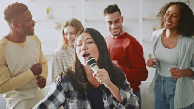 A woman with long dark hair sings into a microphone while friends stand around her, clapping and smiling. The group appears to be having fun during a weekend gathering.