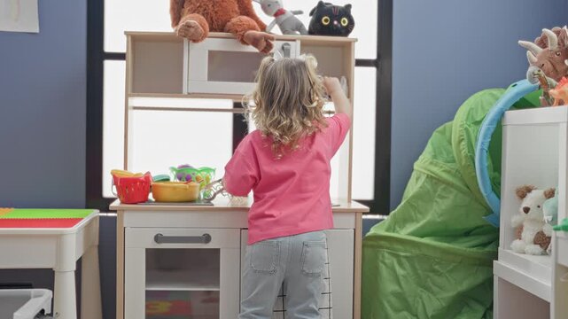 Girl reaches for stuffed bear on wooden play kitchen shelf in a children's playroom inside a building; curiosity.