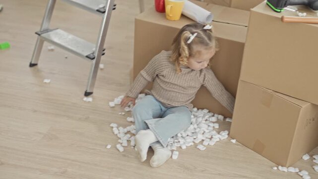 Little girl sitting on packing peanuts, reaching toward cardboard boxes and touching foam pieces near a ladder in a building; curiosity exploration innocence.
