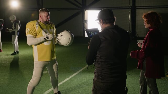 Medium shot of Caucasian female TV show producer explaining shoot idea to muscular American football player, cameraman searching for angle, while working on commercial or documentary at arena
